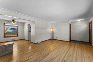 Bright living room with hardwood floors, neutral walls, and a floor lamp. An open doorway leads to a dining area with a ceiling fan and large window. Entrance corridor visible on the right.