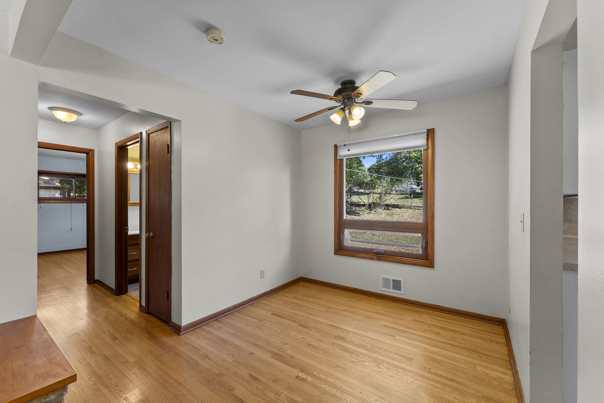 A small empty room with light wood flooring, white walls, a ceiling fan with lights, a window with a view of trees, and doorways leading to a hallway and another room.