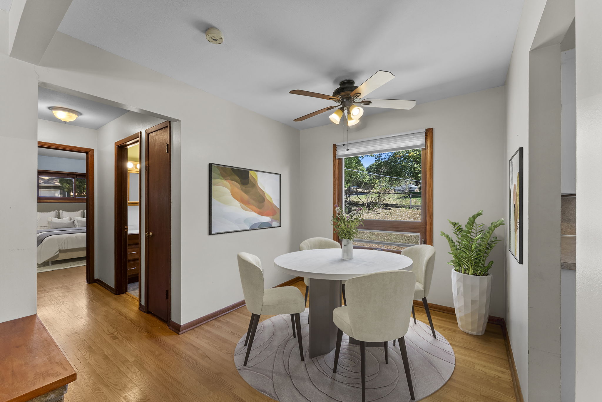 A dining area with a round table and four white chairs sits on a circular rug. A ceiling fan is above, with a window and framed art on the walls. A potted plant is in the corner, and a bedroom is visible through an open doorway.