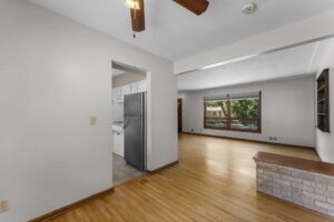 A bright, open living space with hardwood floors, a large window, built-in shelving, and a partial view of a kitchen with white cabinets and a stainless steel refrigerator.