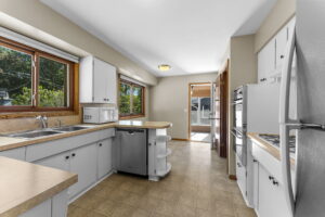 Bright kitchen with white cabinets, beige countertops, double sink by a large window, built-in appliances, tile flooring, and a small island. Sunlight streams in from windows and an open doorway leading to another room.