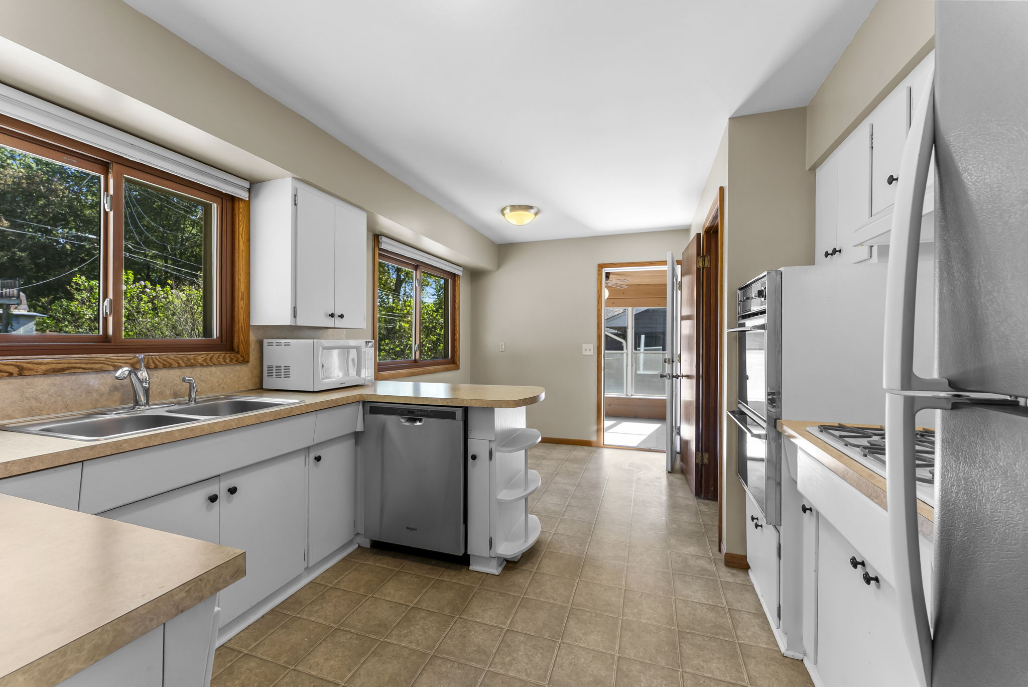 Bright kitchen with white cabinets, beige countertops, double sink by a large window, built-in appliances, tile flooring, and a small island. Sunlight streams in from windows and an open doorway leading to another room.