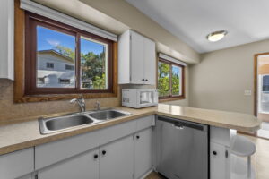 Bright kitchen with white cabinets, a double sink, dishwasher, microwave, and large windows overlooking greenery. There is a breakfast bar with stools and natural light coming in through the windows and door.
