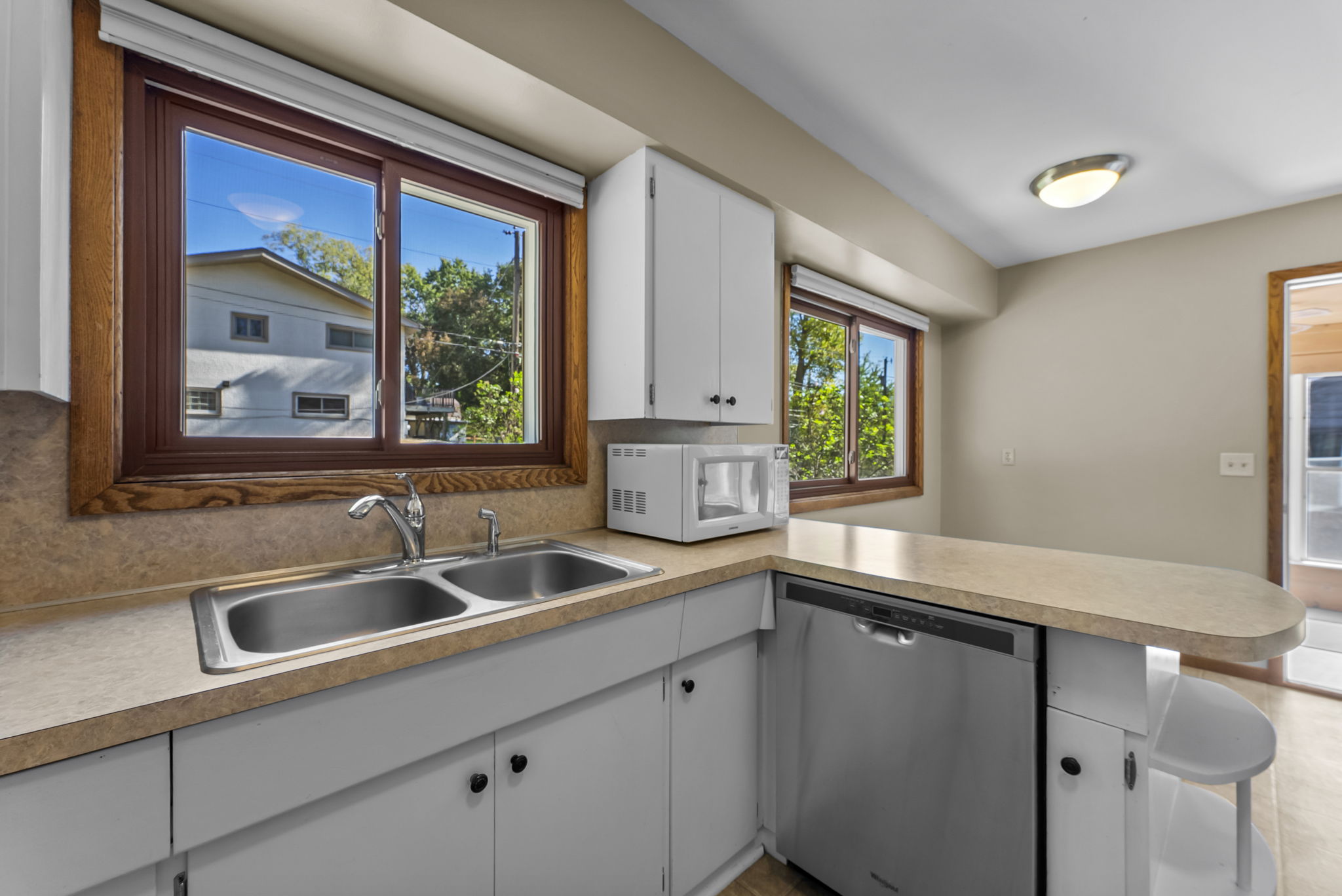 Bright kitchen with white cabinets, a double sink, dishwasher, microwave, and large windows overlooking greenery. There is a breakfast bar with stools and natural light coming in through the windows and door.