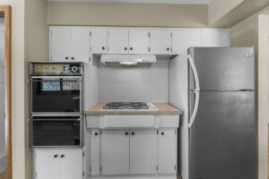 Modern kitchen with white cabinets, a built-in black double oven on the left, a gas cooktop with a range hood in the center, and a stainless steel refrigerator on the right. Neutral wall color and countertop.