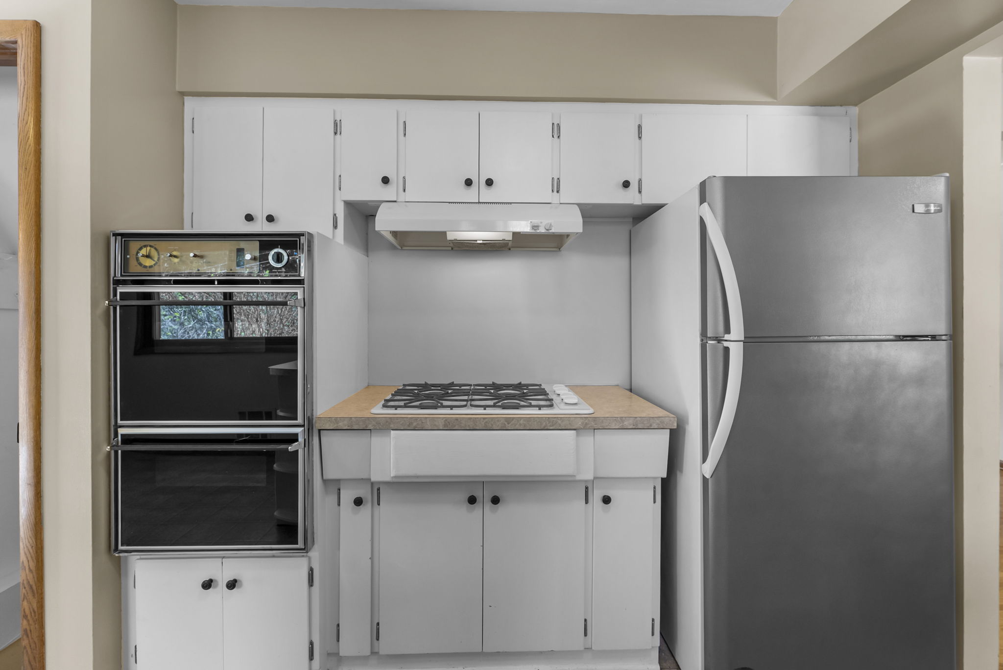Modern kitchen with white cabinets, a built-in black double oven on the left, a gas cooktop with a range hood in the center, and a stainless steel refrigerator on the right. Neutral wall color and countertop.