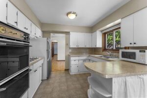 A bright kitchen with white cabinets, beige countertops, a black oven and stove, a microwave, a double sink under a window, and a refrigerator. The floor is tiled and there is a pass-through counter with shelves.