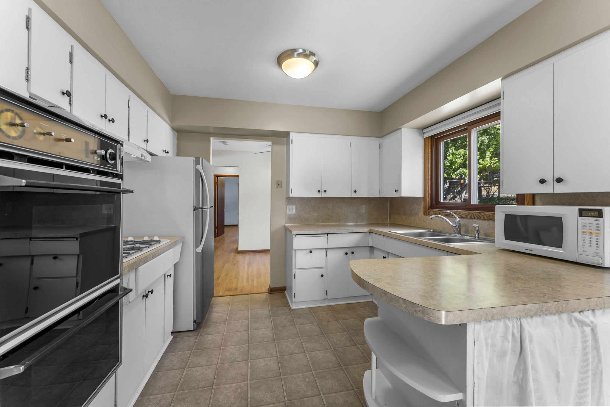A bright kitchen with white cabinets, beige countertops, a black oven and stove, a microwave, a double sink under a window, and a refrigerator. The floor is tiled and there is a pass-through counter with shelves.