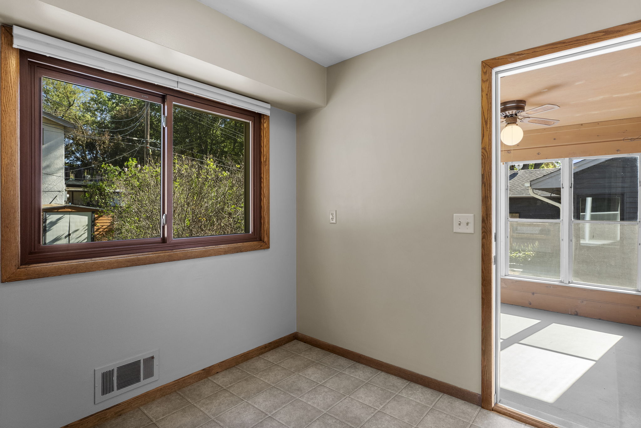A small, empty room with beige walls, a tiled floor, a large window with wood trim, and a door leading to a sunroom with large windows and a ceiling fan. Sunlight brightens both spaces.