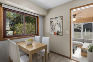 A small dining area with a wooden table, four white upholstered chairs, a bowl of fruit, large window, abstract painting, and a door leading to a covered patio with outdoor seating.