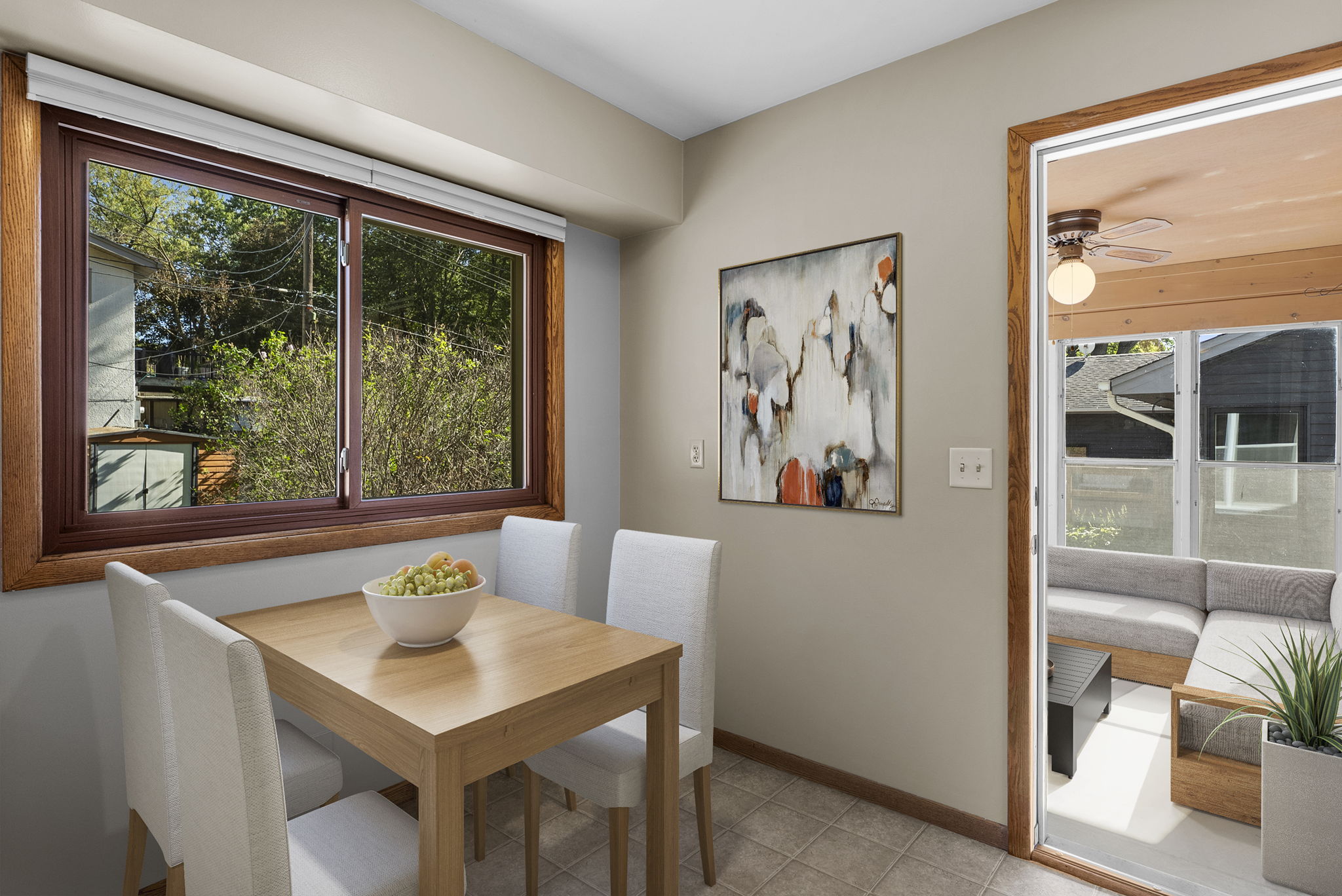 A small dining area with a wooden table, four white upholstered chairs, a bowl of fruit, large window, abstract painting, and a door leading to a covered patio with outdoor seating.