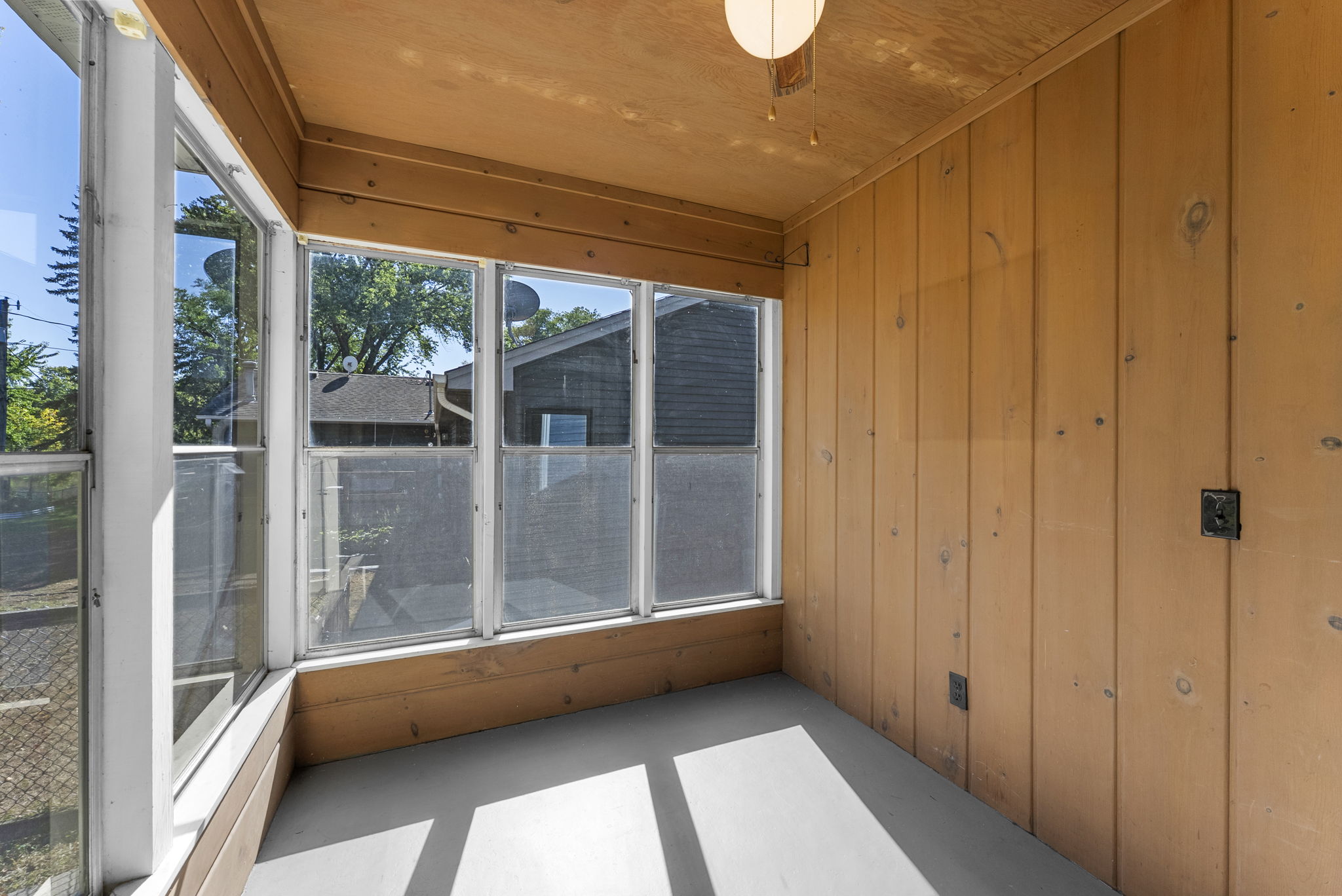 A small sunroom with light wood-paneled walls, large windows letting in natural light, a white ceiling light, and a simple concrete floor. The view outside shows trees, a fence, and neighboring houses.