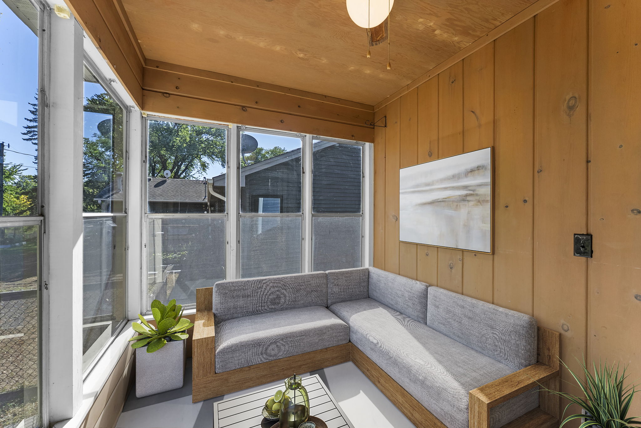 A cozy sunroom with wood-paneled walls, large windows, a modern gray sectional sofa, a coffee table with decorative items, a potted plant, and a wall art piece. Sunlight fills the bright, inviting space.