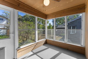 A sunroom with large windows on three sides, a ceiling fan, and a white floor, offering a bright view of trees and neighboring houses outside.