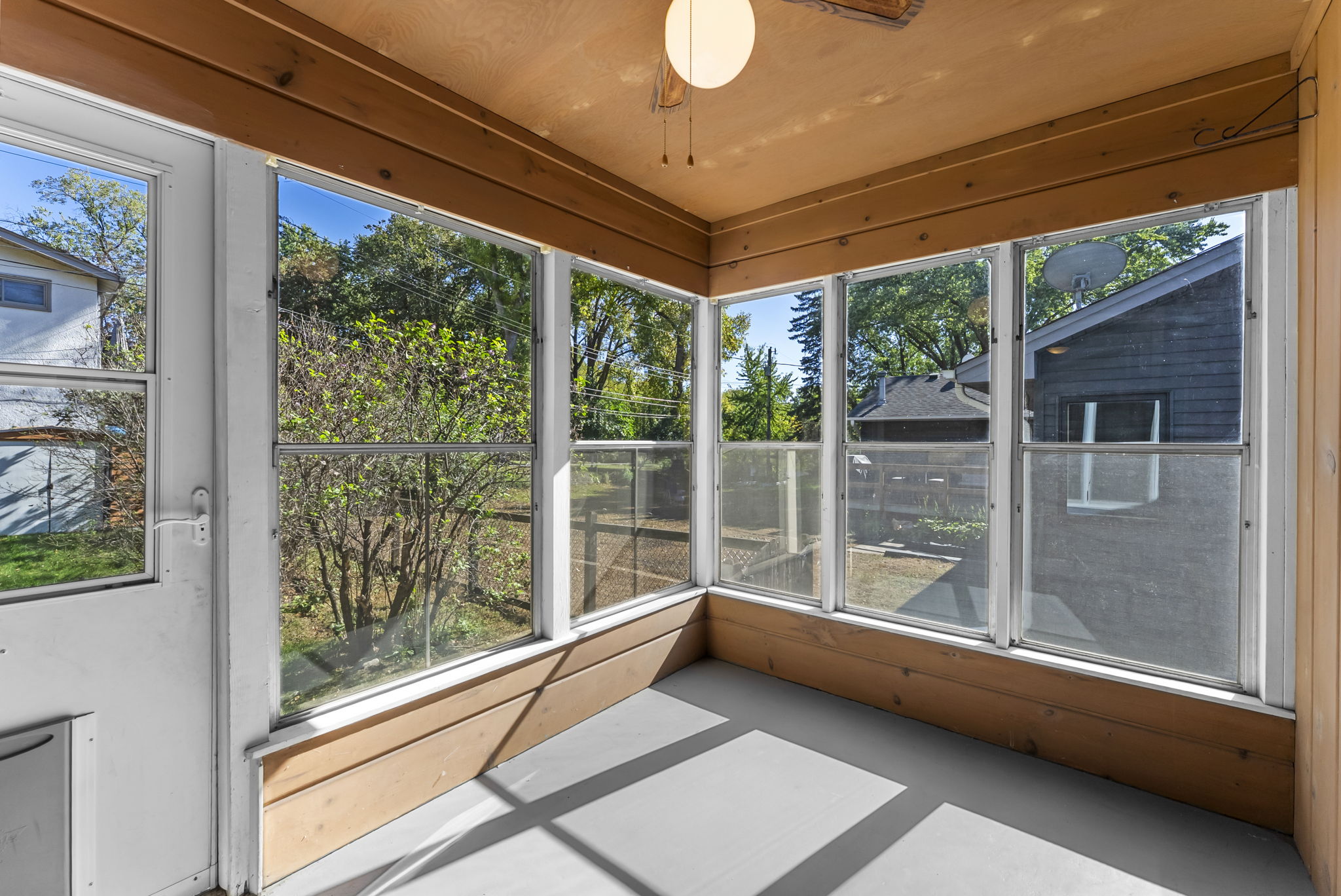 A sunroom with large windows on three sides, a ceiling fan, and a white floor, offering a bright view of trees and neighboring houses outside.