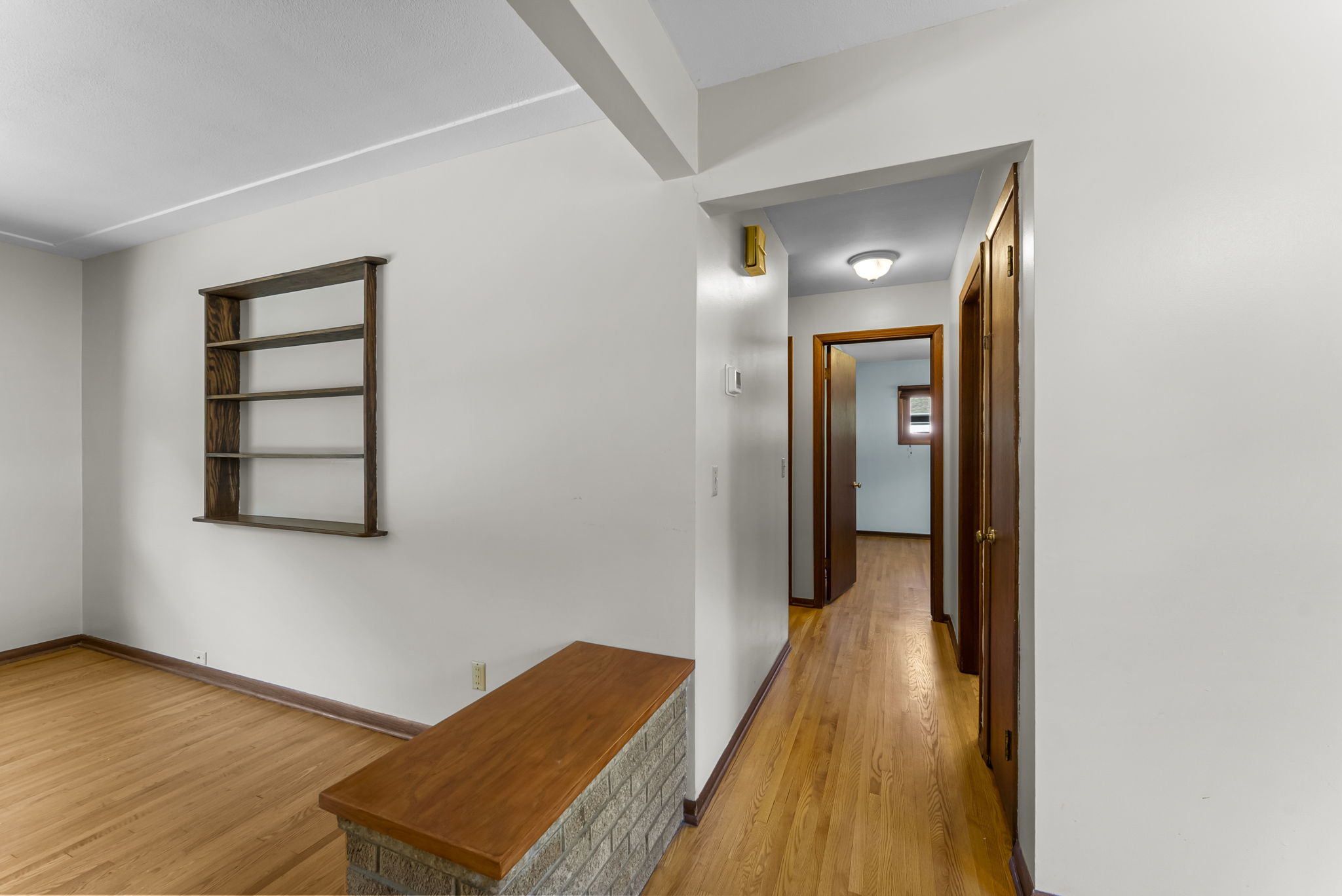 A hallway with wood floors leads to rooms in a house. The walls are white, and a built-in wooden shelf is on the left. A wooden door and a doorway are visible, and a small brick-topped divider is in the foreground.