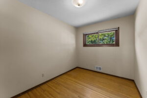 Small empty room with light-colored walls, hardwood flooring, a ceiling light, and a rectangular window with wooden trim, showing green trees outside. The room also has a wall vent and electrical outlets.