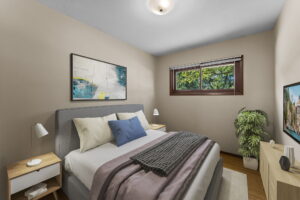 A cozy bedroom with a gray bed, blue accent pillow, two wooden nightstands with lamps, a potted plant, wall art above the bed, a window with a view of trees, and a TV on a cabinet.