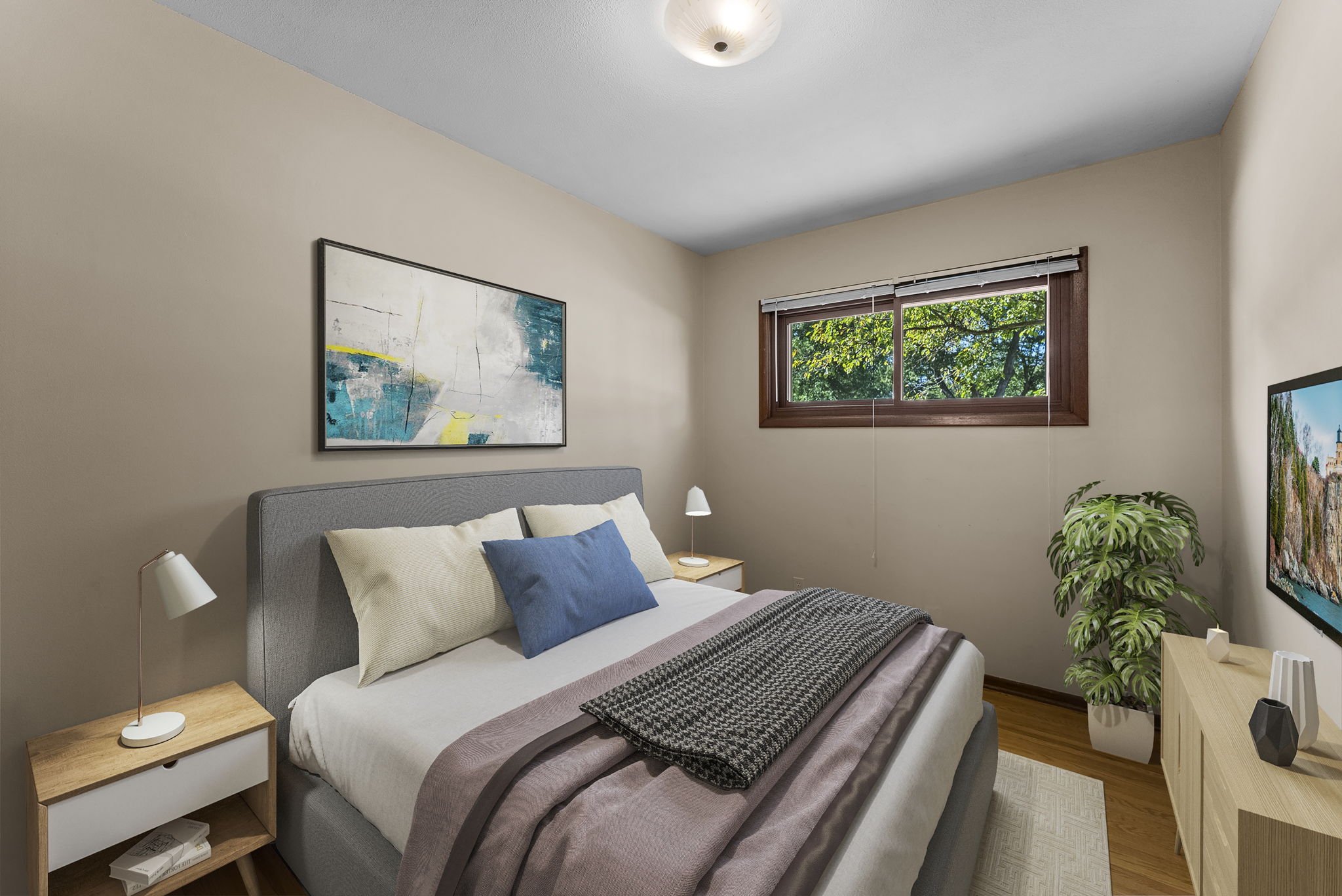 A cozy bedroom with a gray bed, blue accent pillow, two wooden nightstands with lamps, a potted plant, wall art above the bed, a window with a view of trees, and a TV on a cabinet.