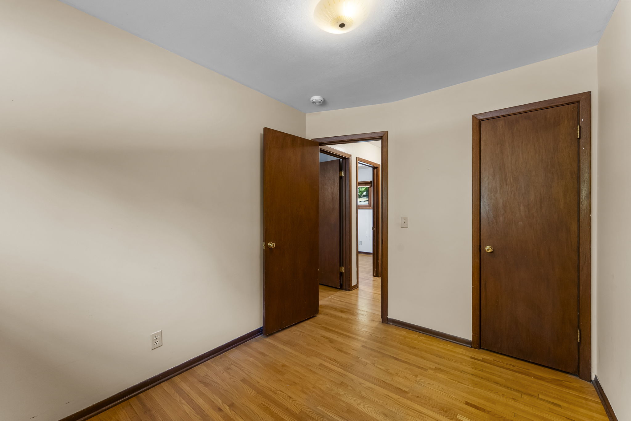 Empty room with light-colored walls, hardwood floor, overhead light, and three wooden doors—two closed and one open, revealing a hallway and another room in the background.