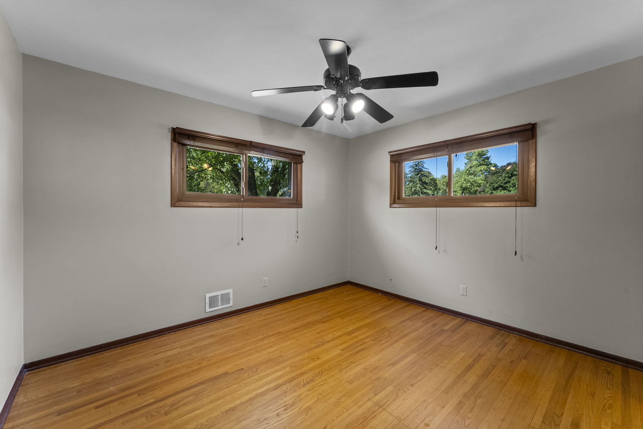 Empty room with light-colored walls, hardwood floors, two rectangular windows with wooden trim, and a ceiling fan with lights. Green trees and blue sky are visible through the windows.