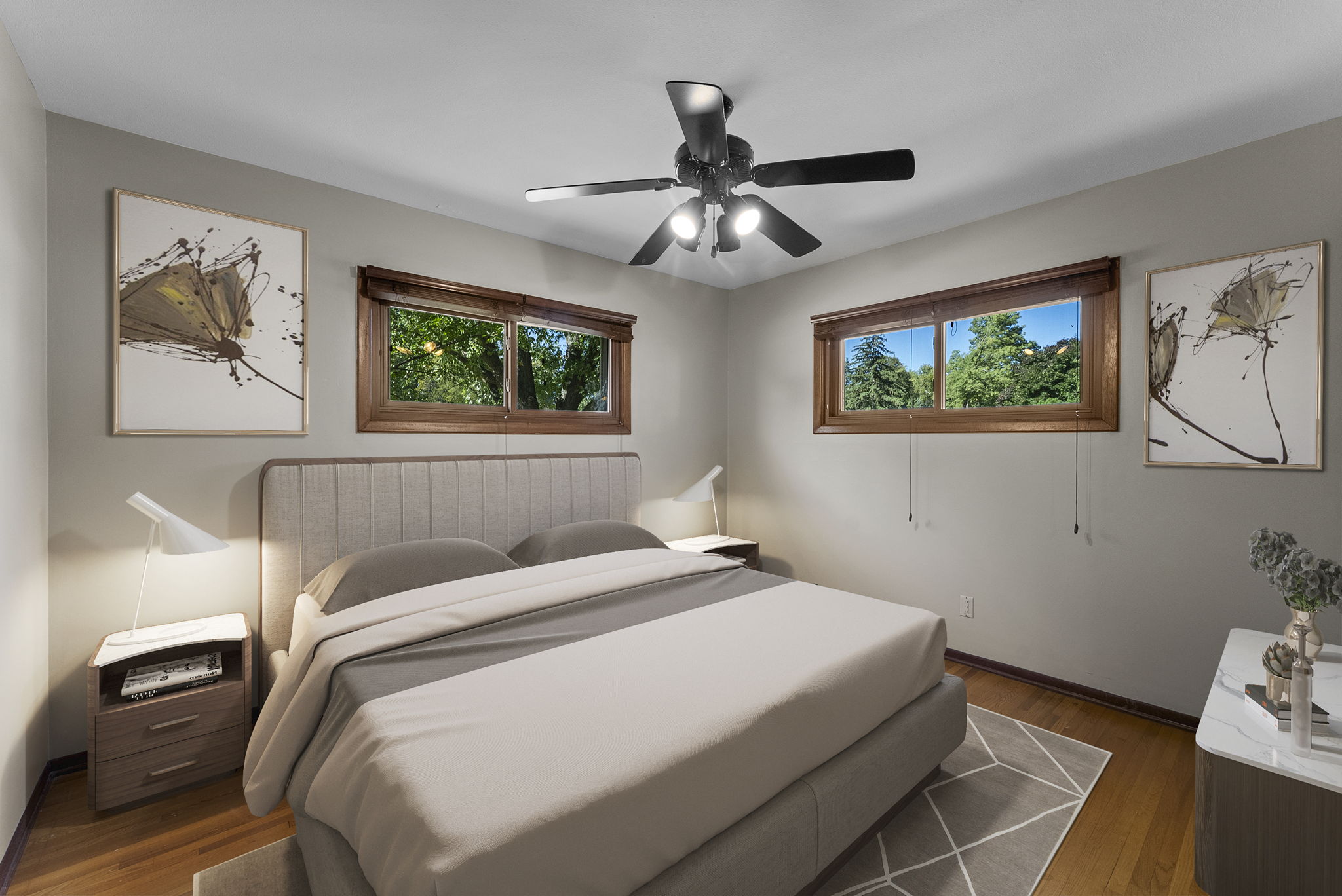 A modern bedroom with a large bed, neutral bedding, wooden floors, two bedside tables with lamps, two windows with wooden frames, nature-themed wall art, and a ceiling fan with lights.