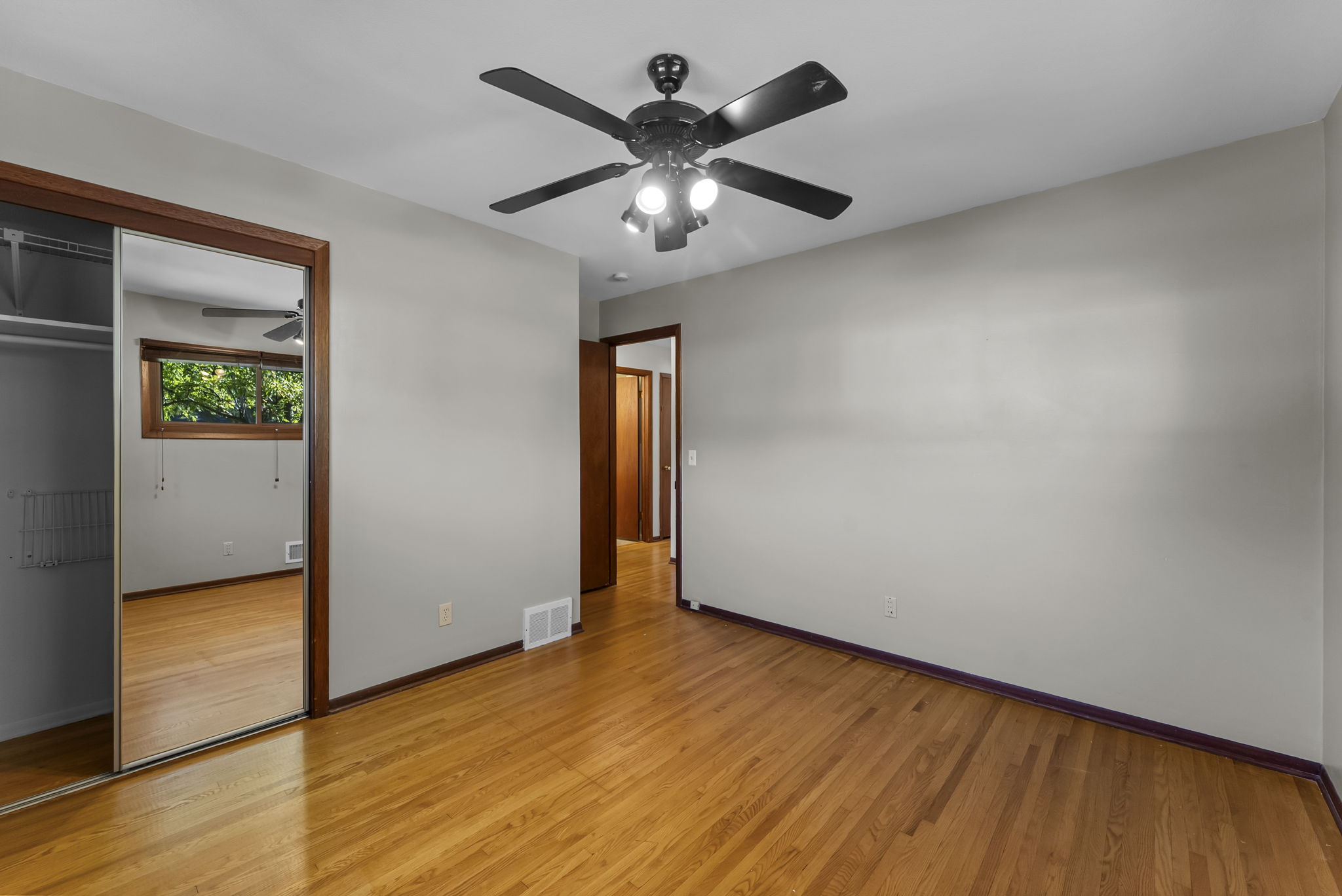 An empty bedroom with light gray walls, wood flooring, a ceiling fan with lights, a mirrored closet door, and a window reflecting greenery outside.