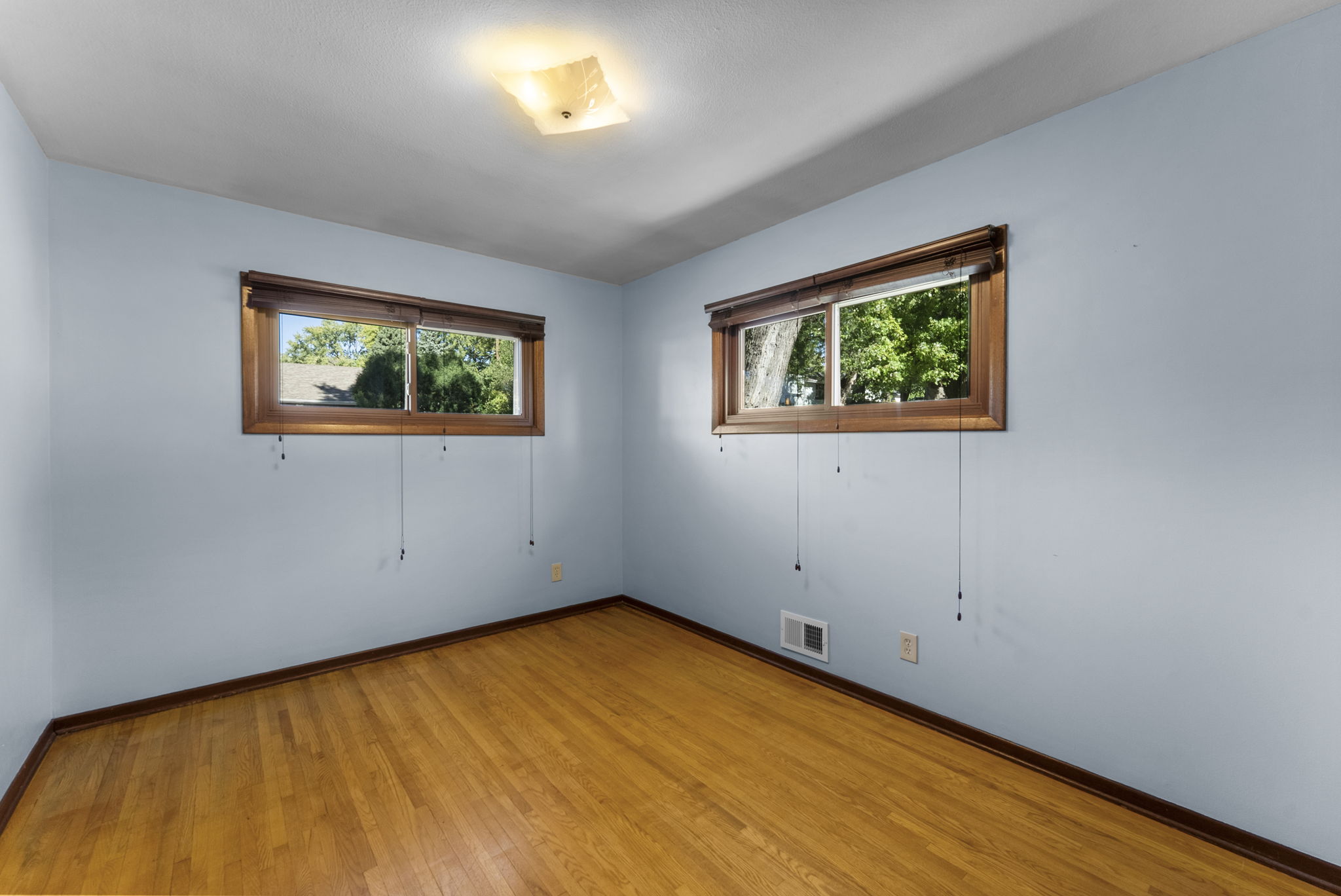 Empty room with light blue walls, hardwood floor, and two rectangular windows with wooden trim. Sunlight is streaming in, and green trees are visible outside the windows. Ceiling light fixture and wall vent are present.