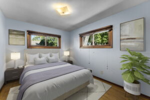 A bedroom with light blue walls, two windows with wooden frames, a double bed with gray and white bedding, two bedside tables with lamps, two abstract paintings, and a potted plant in the corner.