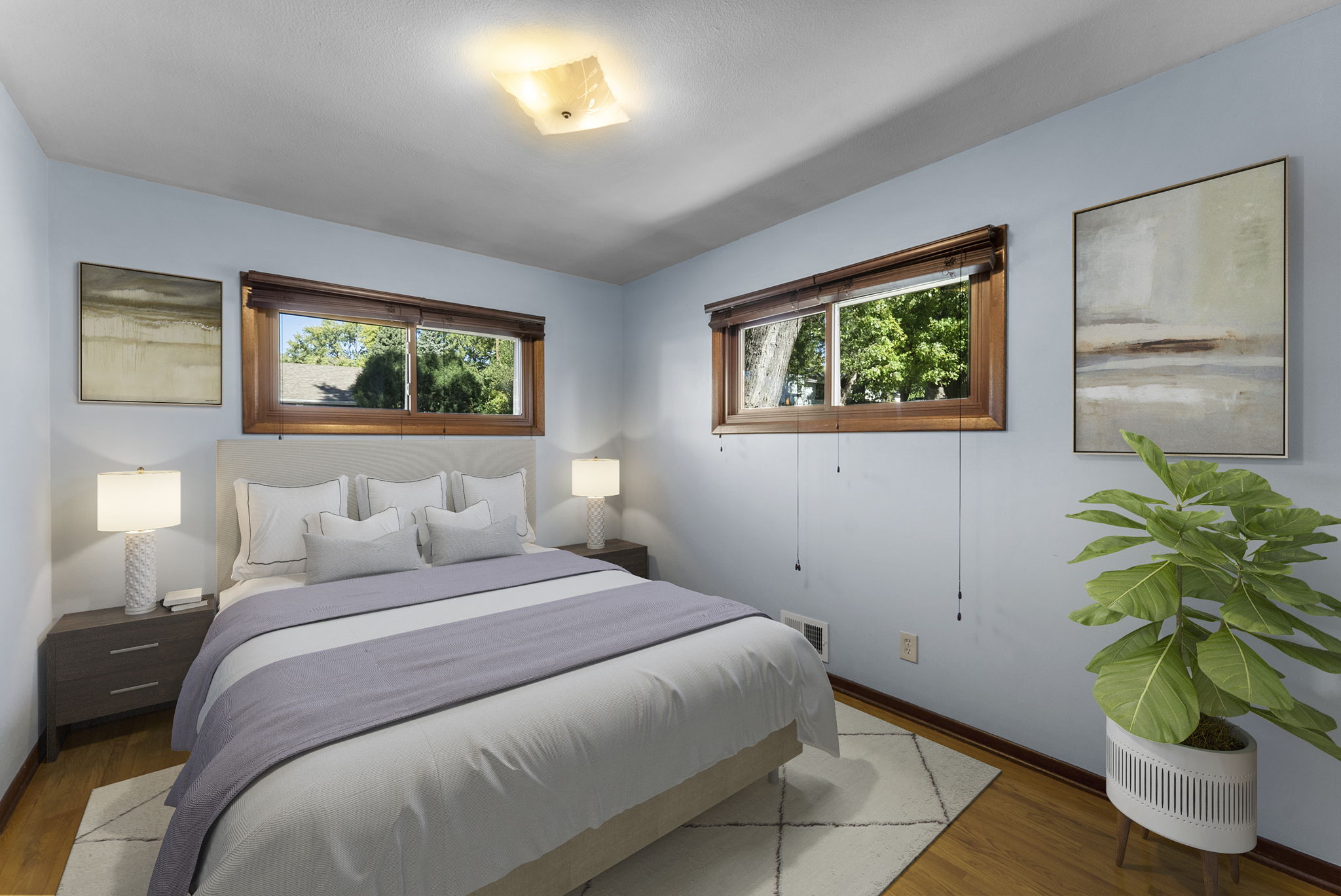 A bedroom with light blue walls, two windows with wooden frames, a double bed with gray and white bedding, two bedside tables with lamps, two abstract paintings, and a potted plant in the corner.