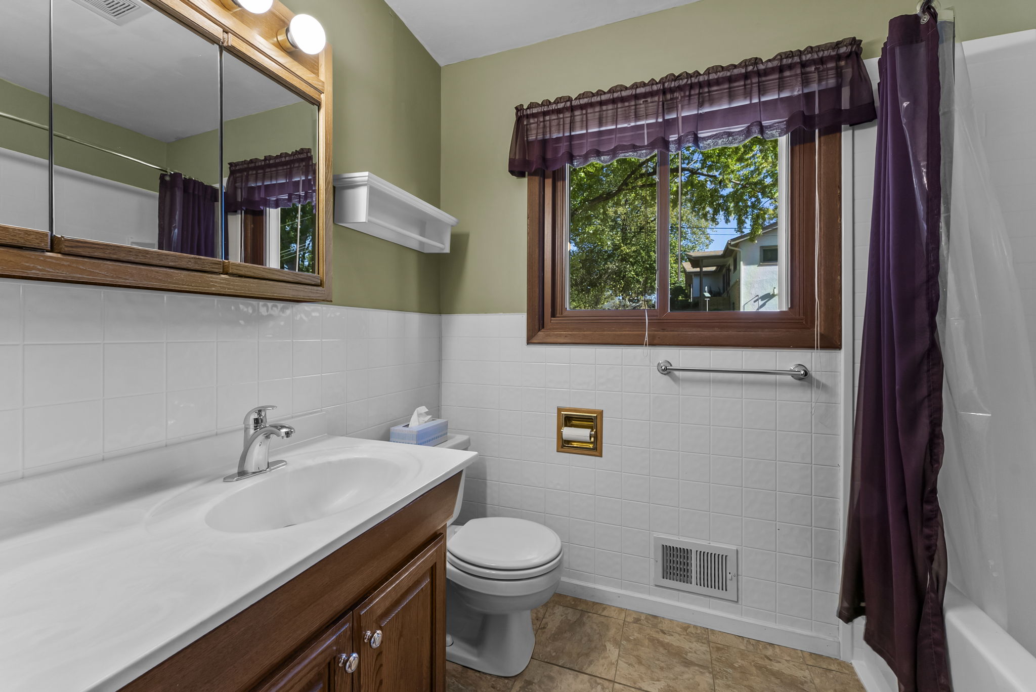 A small bathroom with a vanity sink, mirror cabinet, toilet, and a window with purple curtains. The walls are half white tile and half green paint. There is a bathtub with a purple curtain on the right.