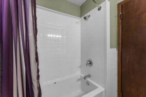 A white bathtub with a tile shower surround, silver faucet, and showerhead. A purple shower curtain is partially closed, and a wooden door is visible to the right. The wall is painted a light green color.
