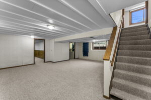 Spacious basement with carpeted floors, white paneled ceiling, stairs with a wooden handrail, small windows for natural light, and multiple rooms visible in the background.