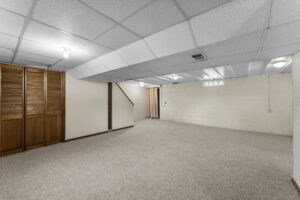 Spacious finished basement with beige carpet, white paneled ceiling, wood closet doors on the left, cinder block walls, and a staircase leading upstairs. Room is empty and well-lit with ceiling lights.
