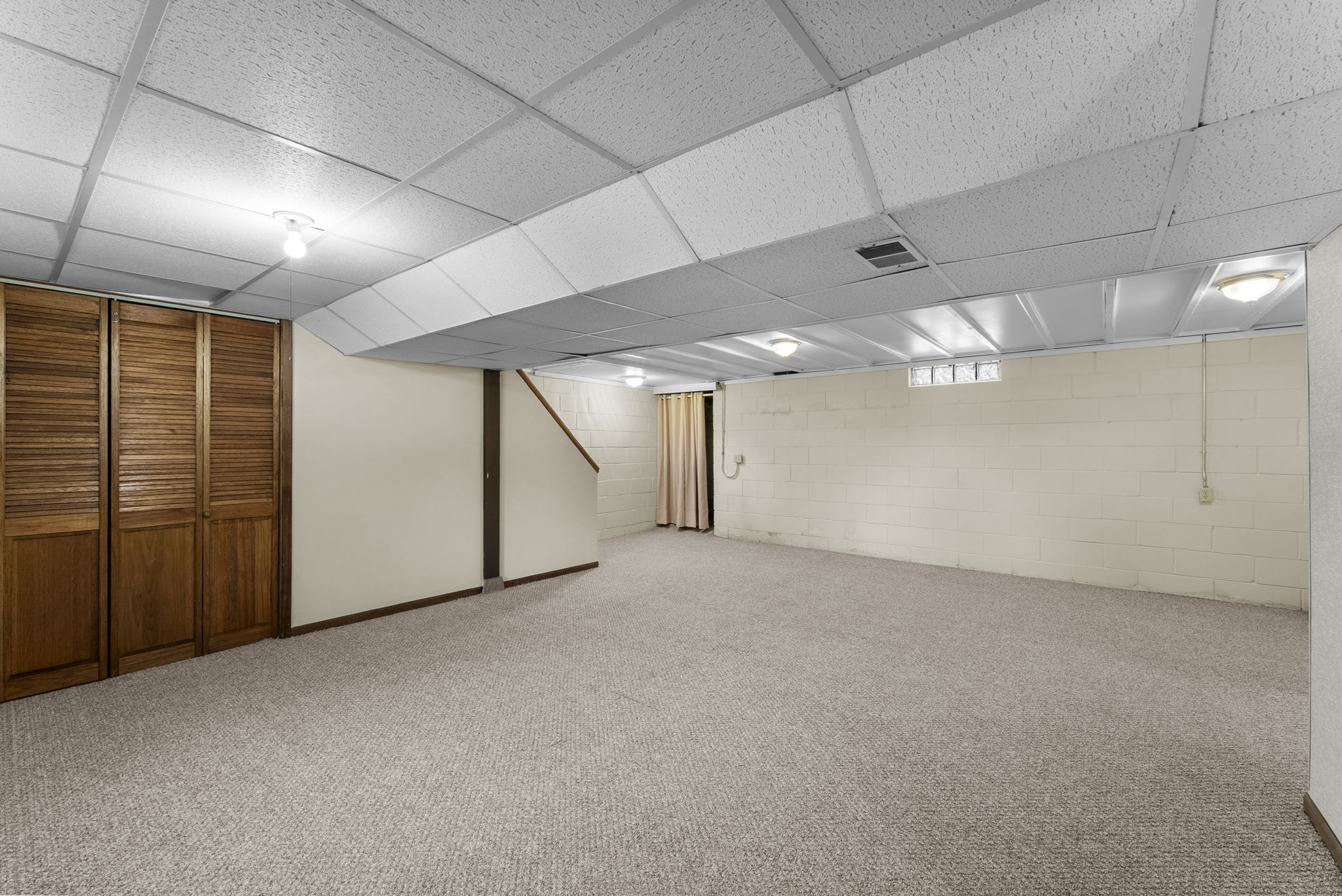 Spacious finished basement with beige carpet, white paneled ceiling, wood closet doors on the left, cinder block walls, and a staircase leading upstairs. Room is empty and well-lit with ceiling lights.
