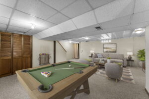 A finished basement with a pool table, cue rack, two gray armchairs, a sofa, a rug, a coffee table, lamps, a potted plant, wall art, and wood-paneled closet doors. Stairs lead up from the room.