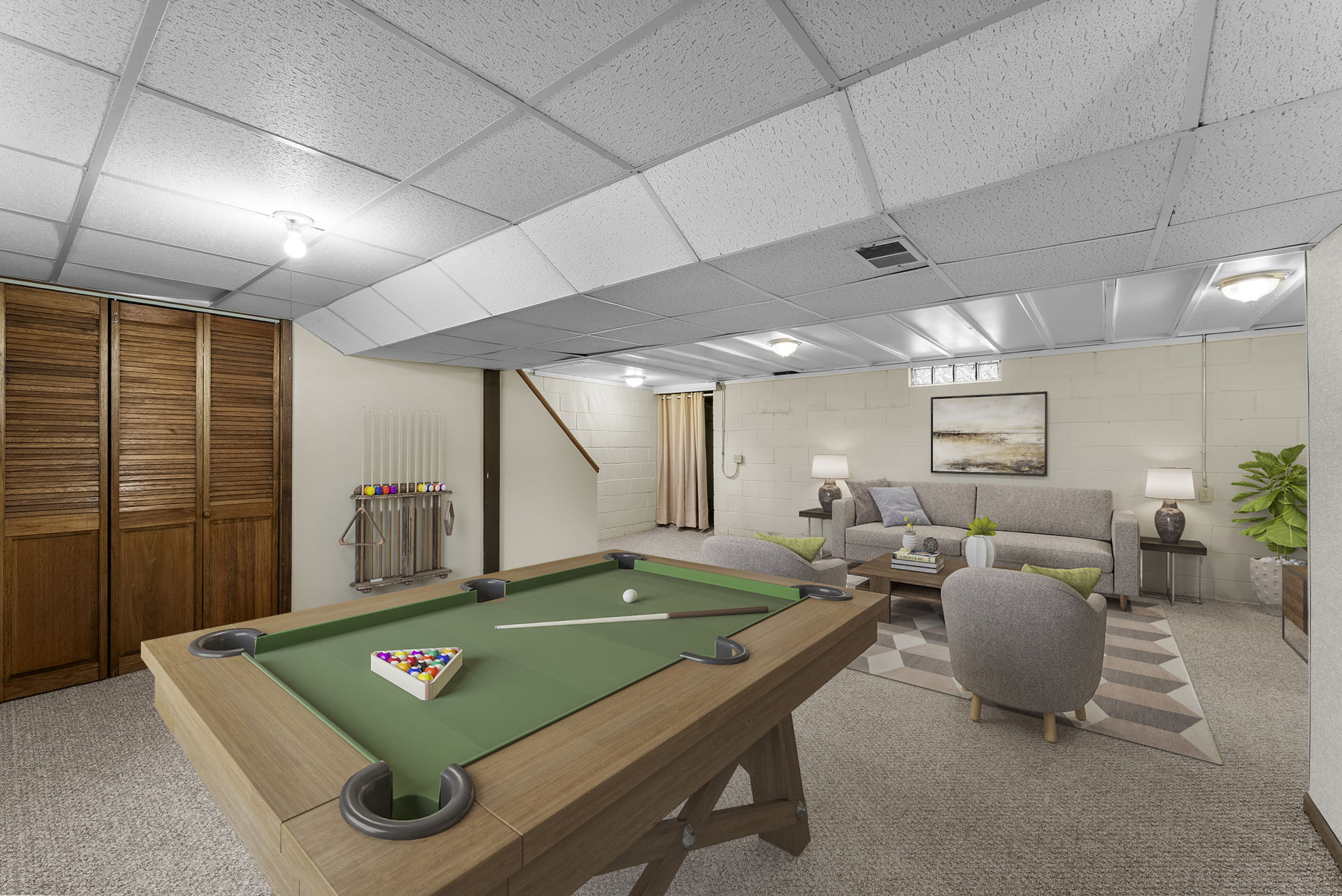 A finished basement with a pool table, cue rack, two gray armchairs, a sofa, a rug, a coffee table, lamps, a potted plant, wall art, and wood-paneled closet doors. Stairs lead up from the room.