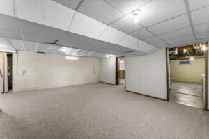 A spacious, empty basement room with beige carpet, white painted cinder block walls, and a drop ceiling with fluorescent lighting. Several doorways lead to unfinished areas with exposed pipes and utilities.