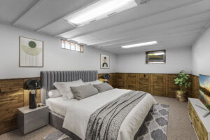 A cozy bedroom with a gray upholstered bed, white bedding, two black lamps on bedside tables, modern artwork on the walls, a TV on a stand, a potted plant, and wood-paneled lower walls with small windows.