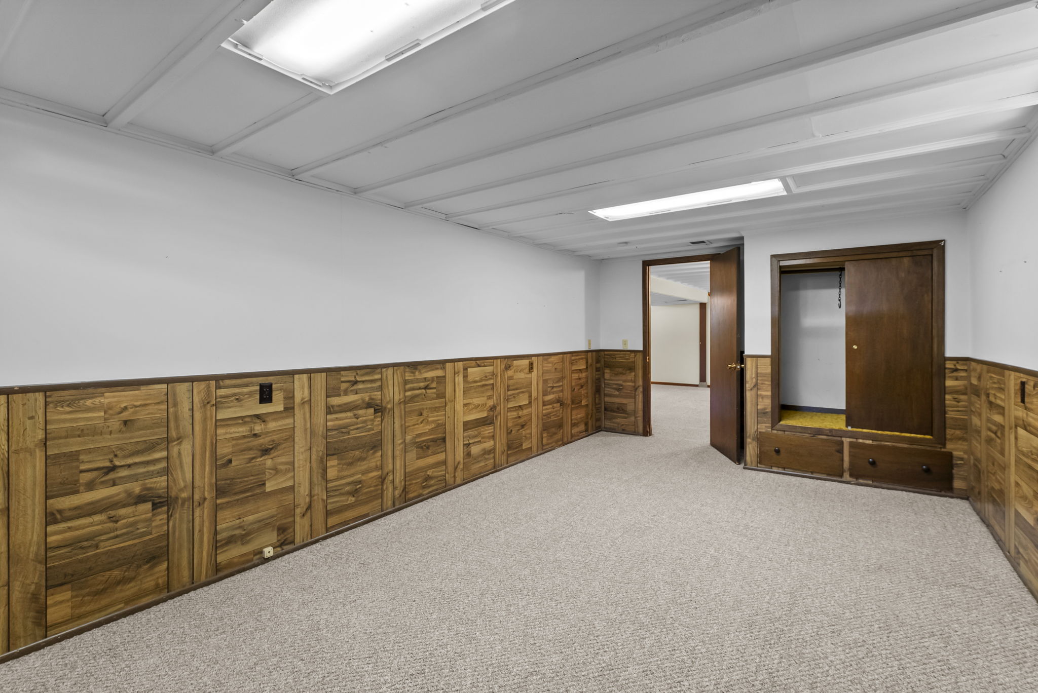 A basement room with wood paneling on the lower half of white walls, beige carpet, a closet with built-in drawers, and an open wooden door leading to another room. Ceiling has fluorescent lighting.