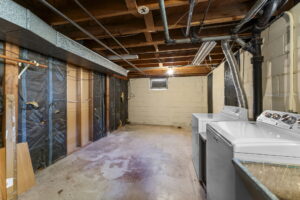 Unfinished basement with exposed beams, pipes, and insulation. There is a washer and dryer on the right side and a small window on the back wall, allowing some natural light into the space.