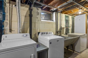 A basement laundry room with exposed pipes and ceiling, a washer and dryer, utility sink, chest freezer, and a small window letting in natural light.