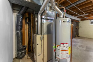 Unfinished basement with an exposed ceiling showing ducts, a gray hot water heater, and a beige furnace unit. The area has concrete floors and a small window letting in natural light.