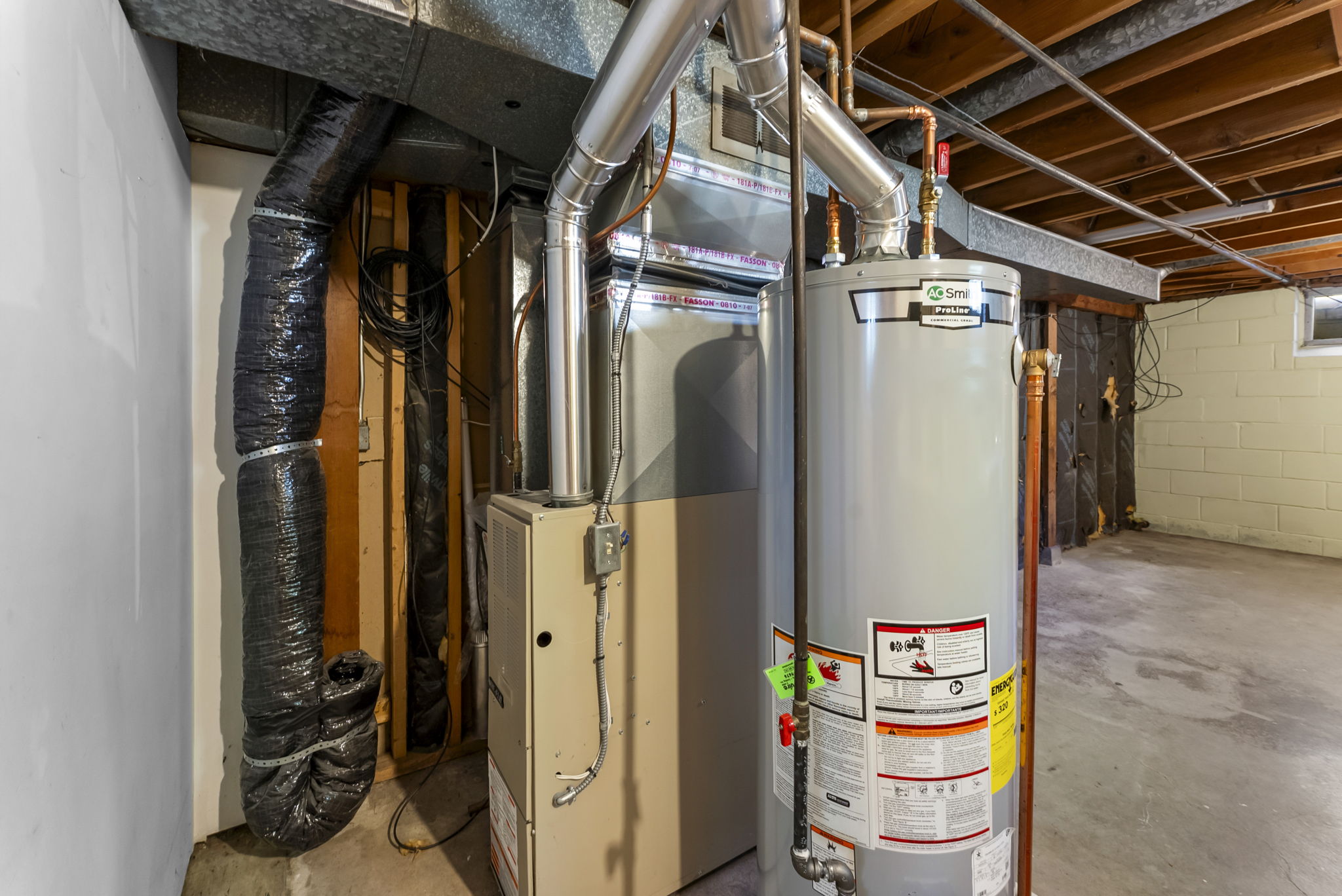 Unfinished basement with an exposed ceiling showing ducts, a gray hot water heater, and a beige furnace unit. The area has concrete floors and a small window letting in natural light.