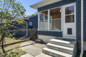 Concrete steps lead to a white door with a dog door, attached to a blue house with large windows. A small fenced yard and a tree are visible on the left side.