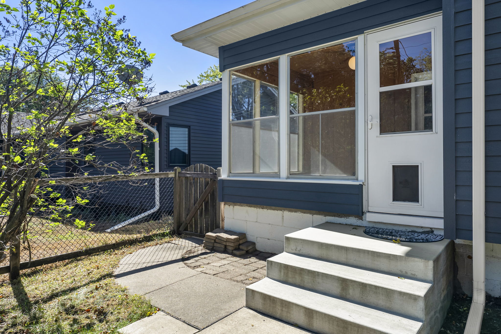 Concrete steps lead to a white door with a dog door, attached to a blue house with large windows. A small fenced yard and a tree are visible on the left side.
