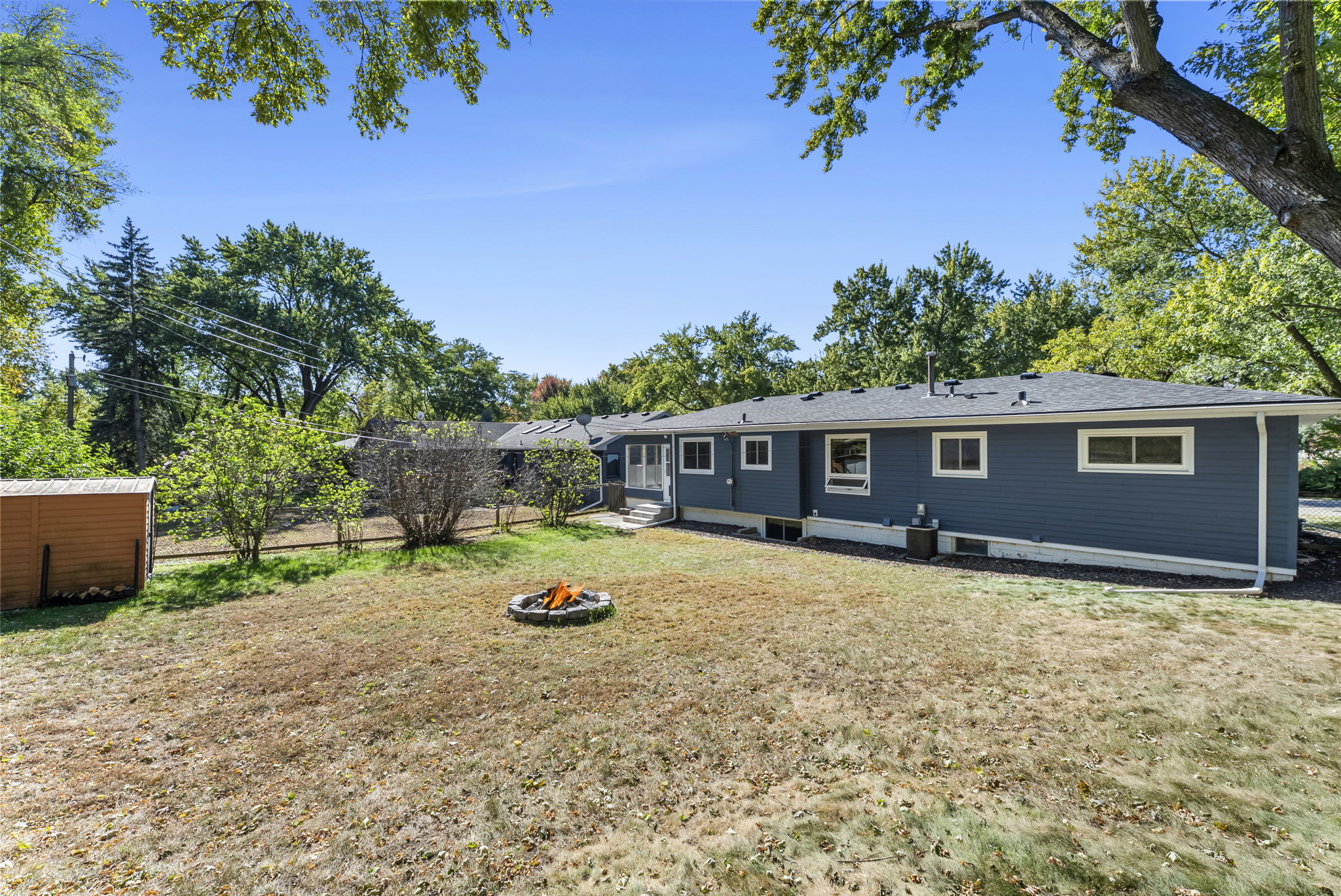 A spacious backyard with a fire pit in the center, a blue single-story house in the background, a small shed to the left, and trees surrounding the yard under a clear blue sky.