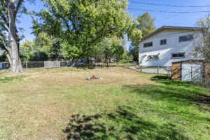 A spacious backyard with green grass, a fire pit in the center, tall trees, a wooden shed, wire fencing, and a white house in the background under a clear blue sky.