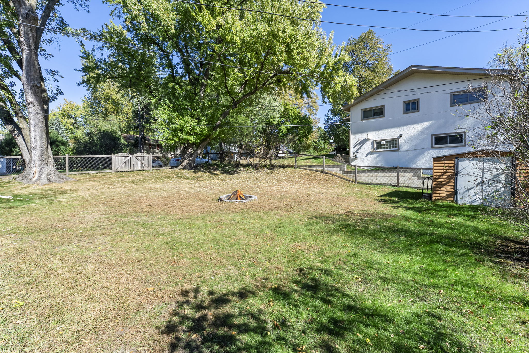 A spacious backyard with green grass, a fire pit in the center, tall trees, a wooden shed, wire fencing, and a white house in the background under a clear blue sky.