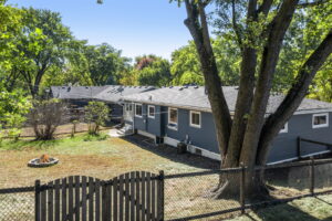 A fenced backyard with a large tree, a fire pit, and a gray single-story house with white trim, set among green trees under a clear blue sky.